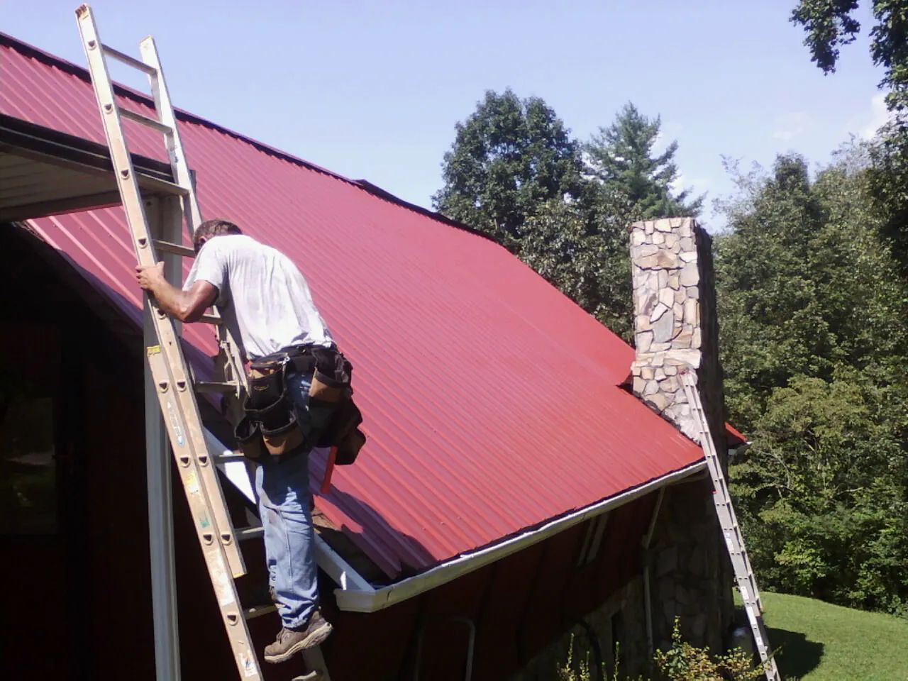Roofer on a ladder beside a red metal roof and stone chimney — Soffit & Fascia Repair by Roofing Brothers in North Washington